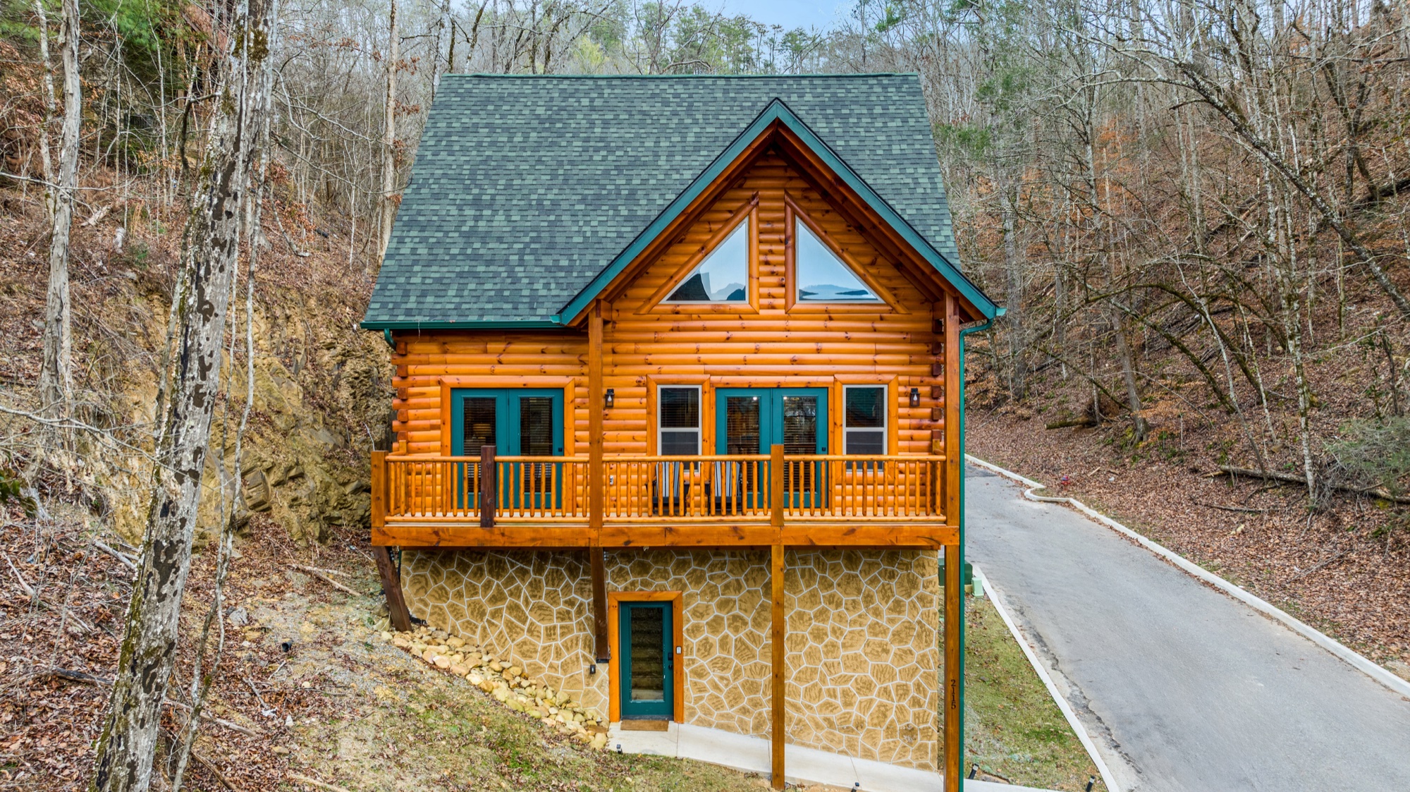 Front view of Whispering Valley Cabin in daylight, with its log balcony, green-trimmed roof, and stone foundation